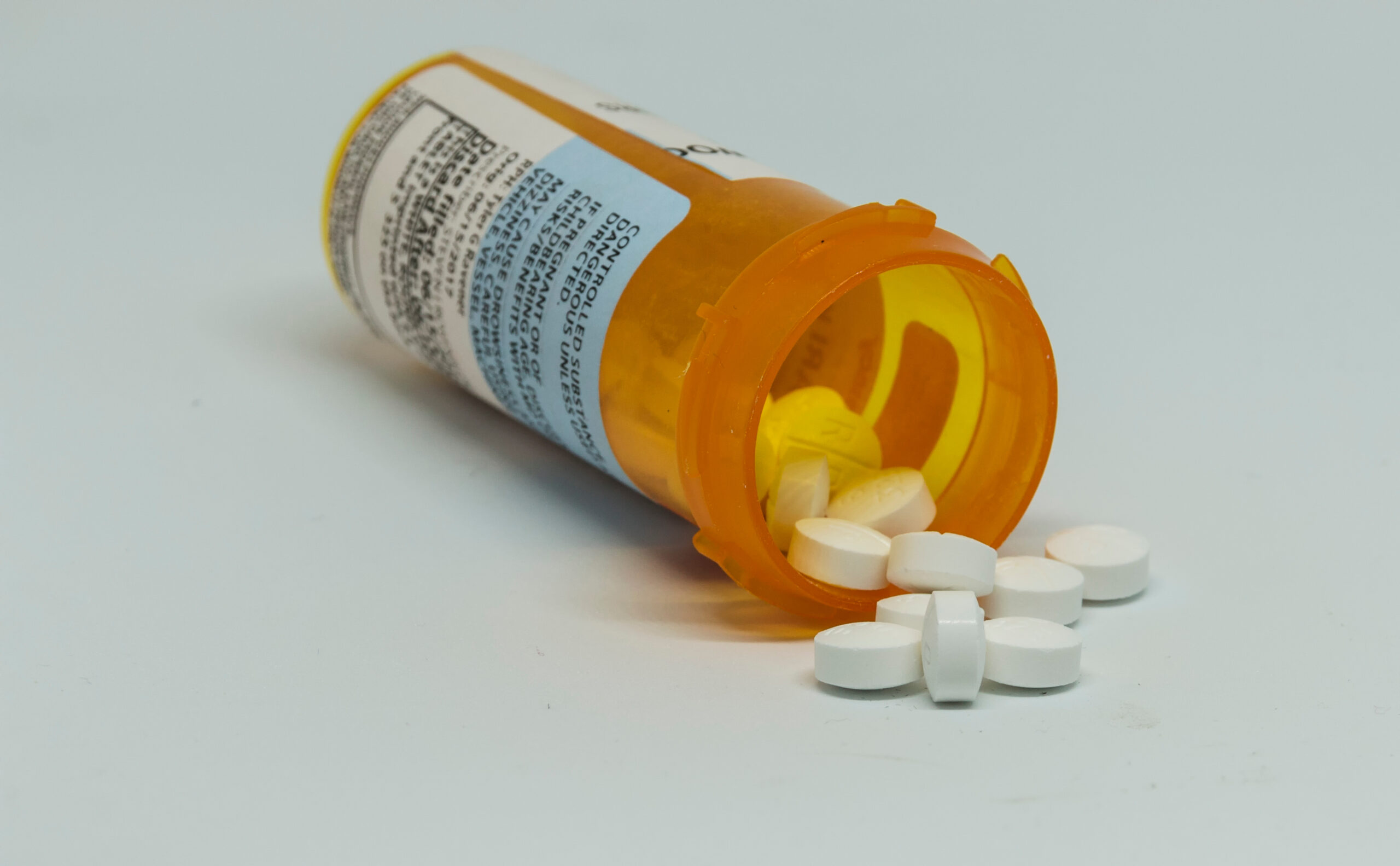 Pill bottle on its side with controlled substance pills falling out of the bottle with a white background.