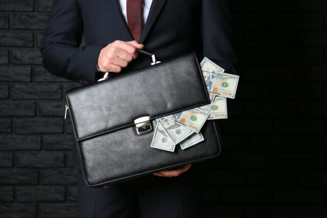 Businessman holding briefcase with dollar banknotes on dark background. Corruption concept