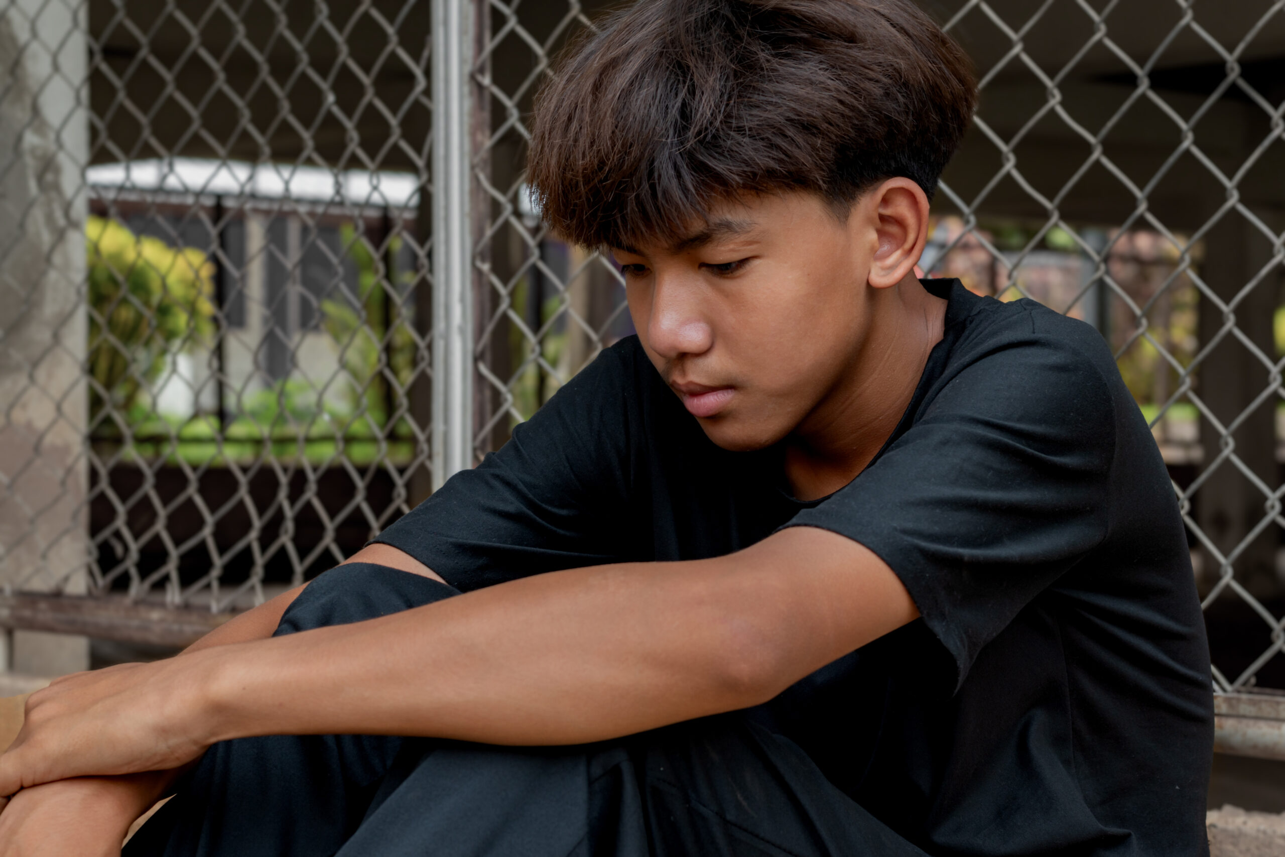 eenboy in a black shirt sits with his knees pressed against a metal fence panel in a juvenile detention facility, awaiting further release, freedom and detention of people concept.